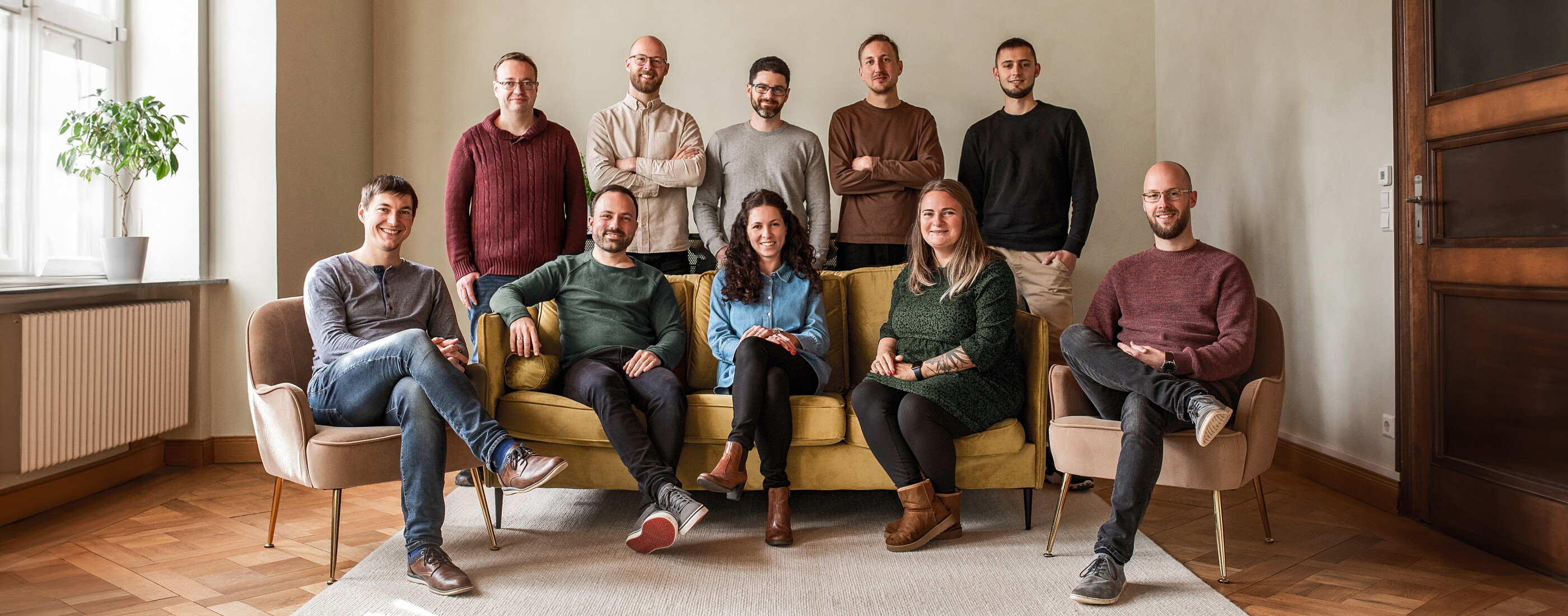 Gruppenfoto des POSTYOU-Teams im Besprechungsraum der Villa: Elf Mitarbeitende sitzen auf einem gelben Sofa und in Sesseln oder stehen dahinter. Der Raum hat Parkett, eine rote Stuckdecke und hohe Fenster.