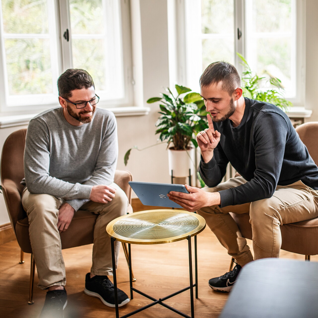 Zwei Mitarbeiter des POSTYOU-Teams sitzen in Sesseln an einem kleinen goldenen Tisch. Einer zeigt auf ein Tablet, der andere schaut lächelnd zu; dahinter ein helles Fenster und Pflanzen.