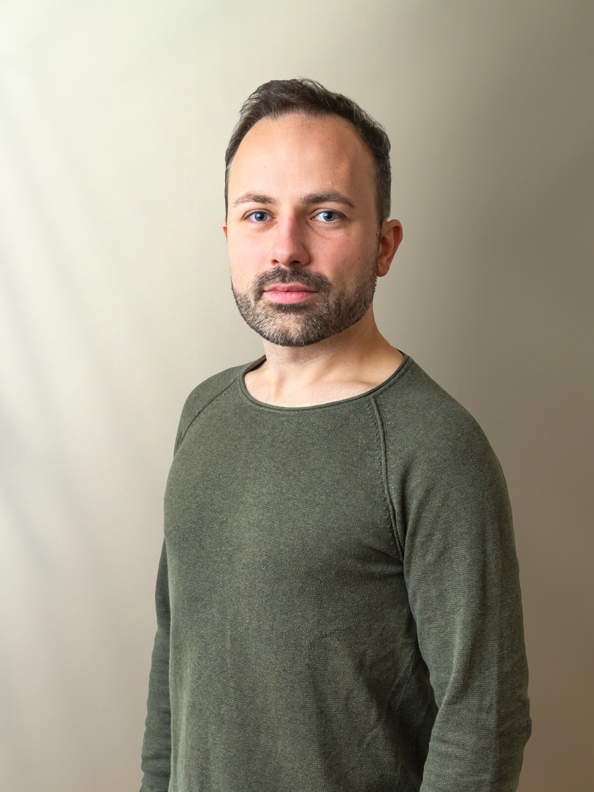 Porträt von Christoph: Er trägt einen olivgrünen Pullover, einen kurzen Bart und blickt ernst in die Kamera. Der Hintergrund ist hell und schlicht.