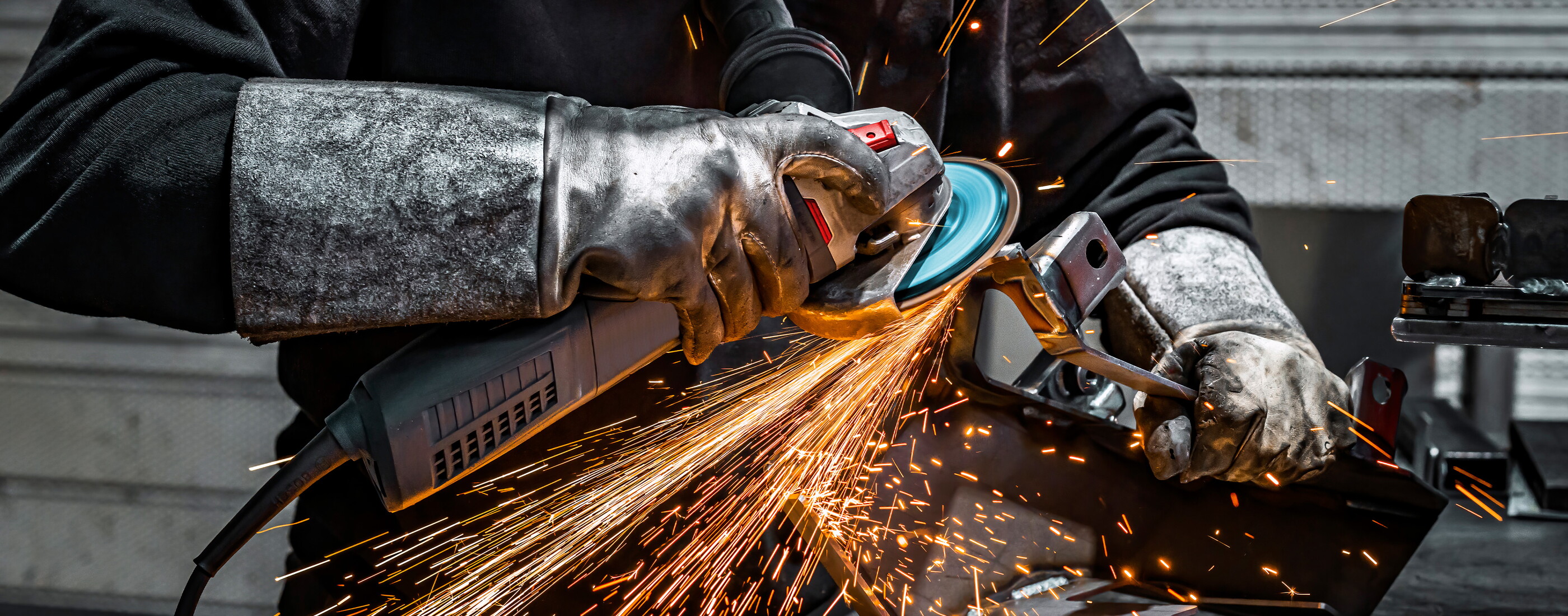 Eine Person mit dicken Schutzhandschuhen schleift mit einem Winkelschleifer an einem Metallteil, ein heller Funkenregen sprüht vor dunklem Hintergrund.