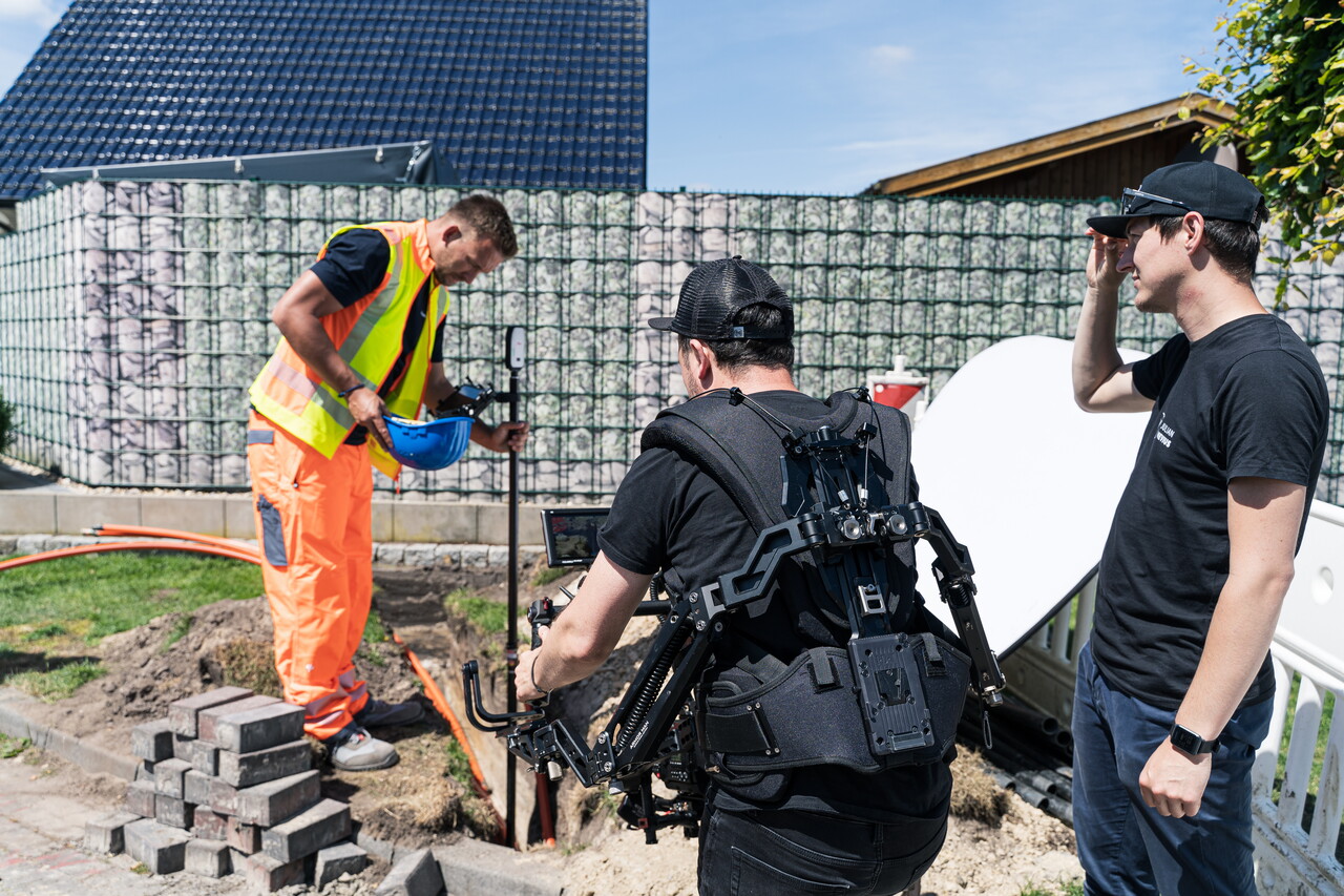Dreh auf einer Baustelle: Ein Bauarbeiter in Warnkleidung arbeitet an einer Grube, ein Kameramann filmt ihn mit einer Kamera an einer Tragweste. Ein weiterer Kollege schaut daneben zu.