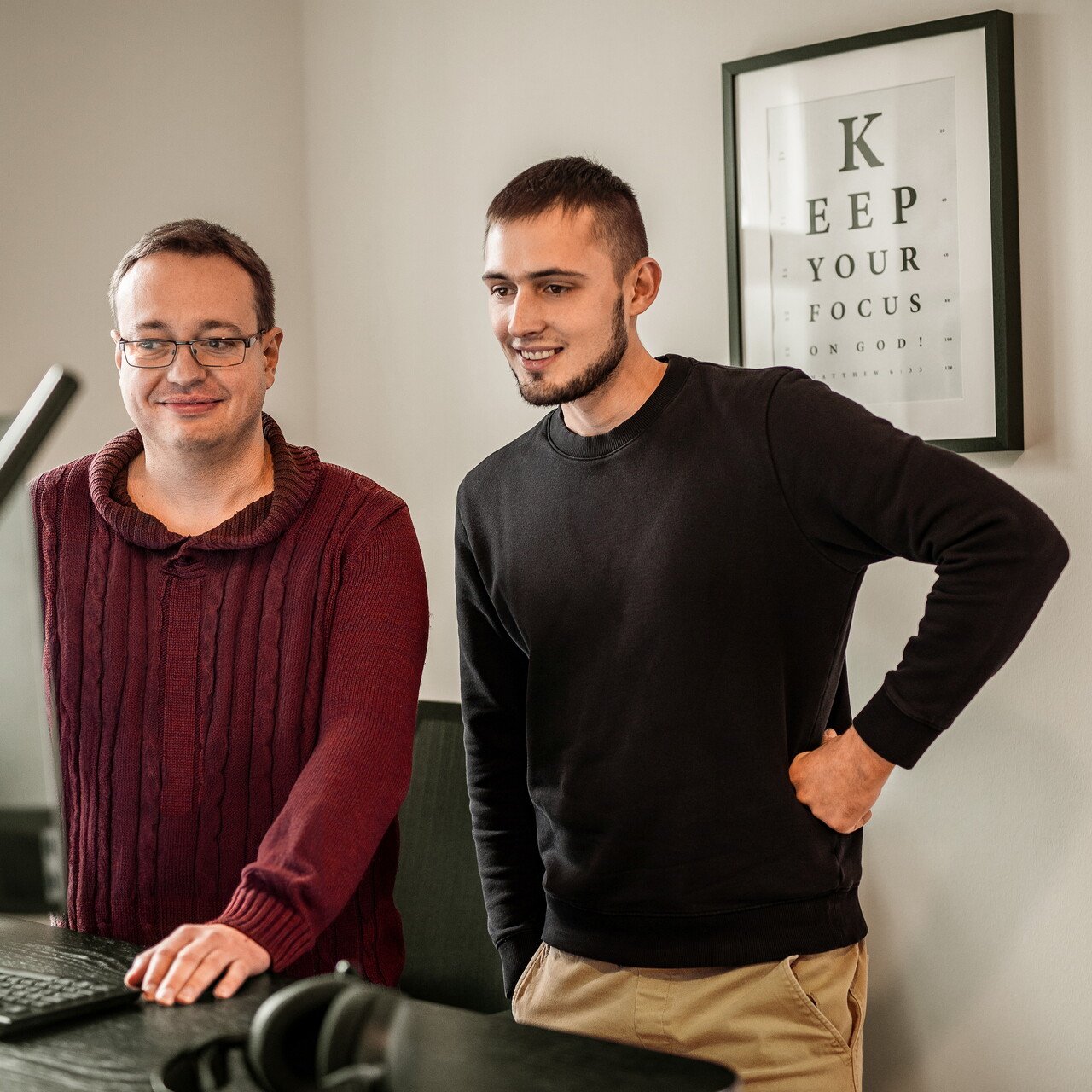 Zwei Mitarbeiter des POSTYOU-Teams lachen gemeinsam an einem Schreibtisch mit Monitor und Tastatur. An der Wand hängt ein Poster im Stil einer Sehtafel mit dem Spruch „Keep your focus“.