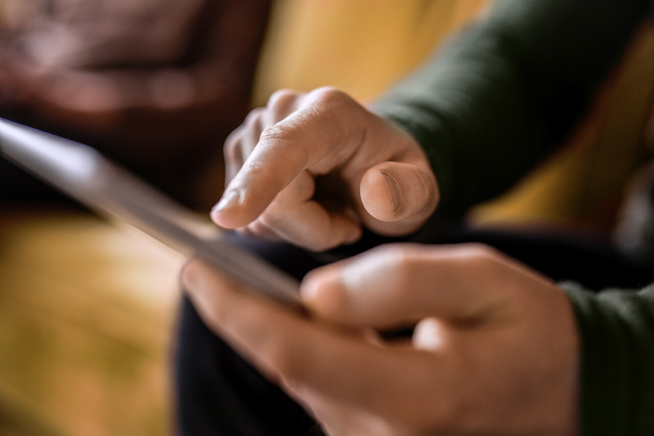 Nahaufnahme: Ein Mitglied des POSTYOU-Teams hält ein Tablet und tippt mit dem Zeigefinger auf den Bildschirm. Der Hintergrund ist warm und unscharf.