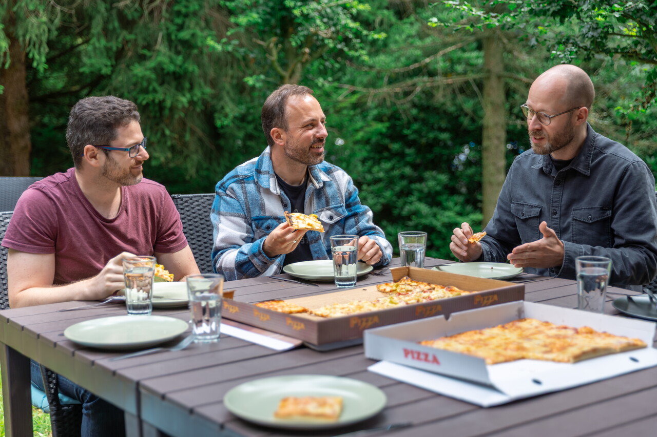 Drei Kollegen sitzen an einem Gartentisch und essen Pizza aus offenen Kartons. Im Hintergrund stehen dichte grüne Bäume.