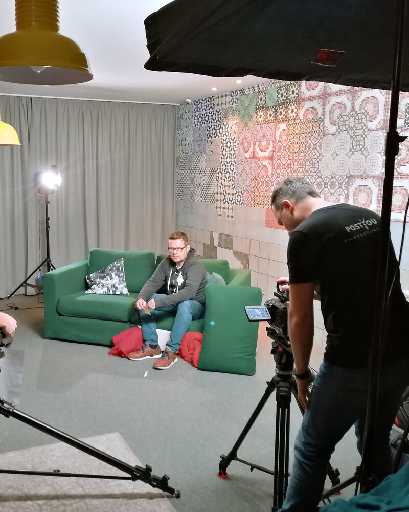 Interviewdreh in einem Studio mit bunter Fliesenwand: Ein Mann sitzt auf einem grünen Sofa, zwei Kameraleute richten ihre Kameras ein. Softboxen und Leuchten stehen ringsum.