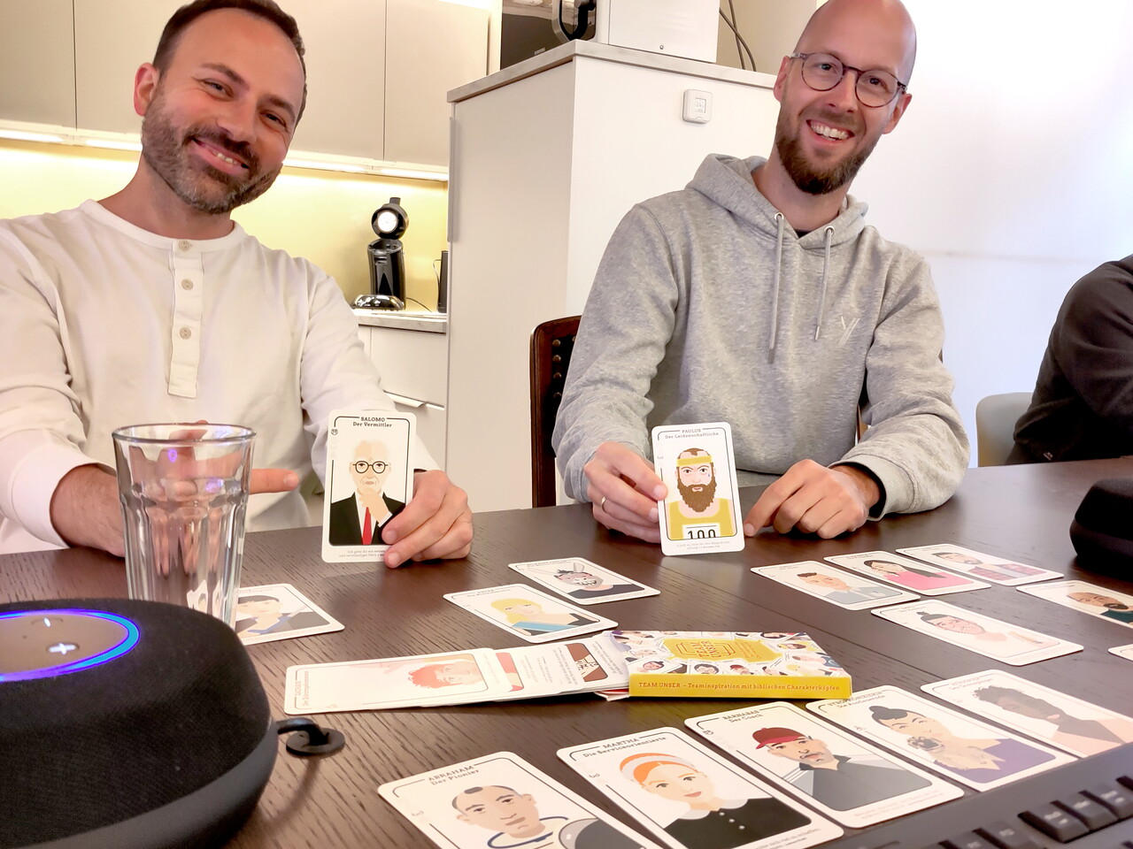 Zwei Kollegen sitzen lächelnd an einem Tisch und halten je eine Spielkarte mit gezeichnetem Gesicht hoch. Vor ihnen liegen weitere Karten mit Porträts ausgebreitet.