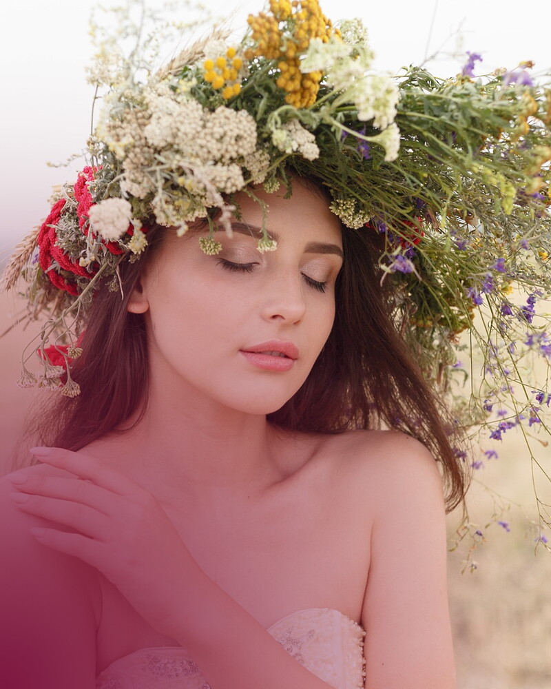 Eine junge Frau mit geschlossenen Augen trägt einen üppigen Kranz aus Wiesenblumen im langen Haar. Das Bild ist in warme, rosa Töne getaucht.