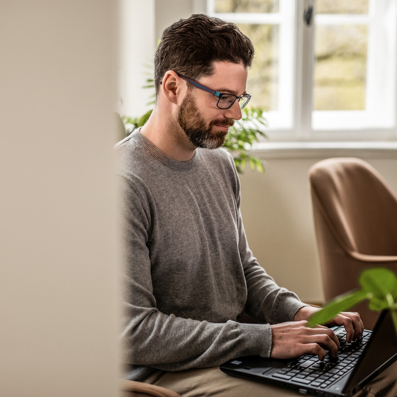 Ein Mitarbeiter des POSTYOU-Teams mit Bart und Brille sitzt konzentriert im Sessel und tippt auf einem Laptop. Im Hintergrund stehen ein weiterer Sessel und ein helles Sprossenfenster.