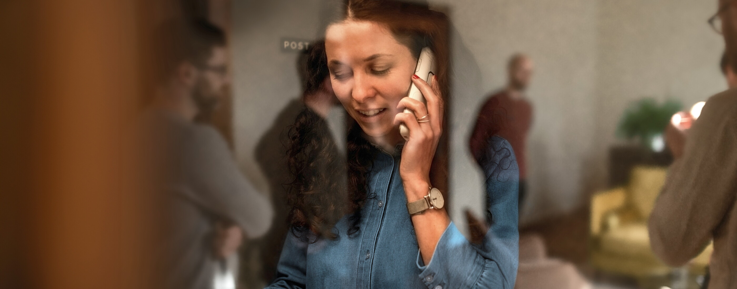 Eine Mitarbeiterin des POSTYOU-Teams im Jeanshemd telefoniert lächelnd mit dem Handy. Sie ist durch eine Glasscheibe fotografiert, in der sich Kollegen und der Büroraum spiegeln.