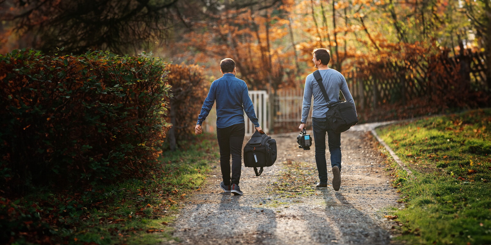 Zwei Mitarbeiter des POSTYOU-Teams gehen, von hinten gesehen, einen Kiesweg entlang und tragen Taschen mit Filmausrüstung. Der Weg ist von Hecken und herbstlich gefärbten Bäumen gesäumt.