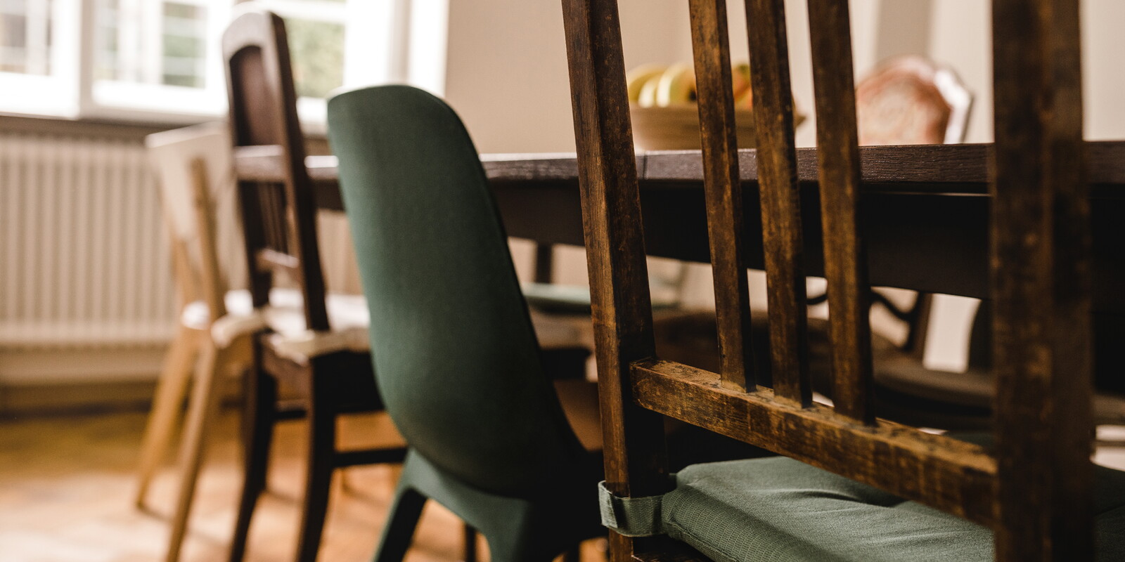 Blick durch die Lehne eines alten Holzstuhls auf den langen Esstisch der Agentur. Um den Tisch stehen verschiedene Stühle, darunter ein grüner Schalenstuhl.
