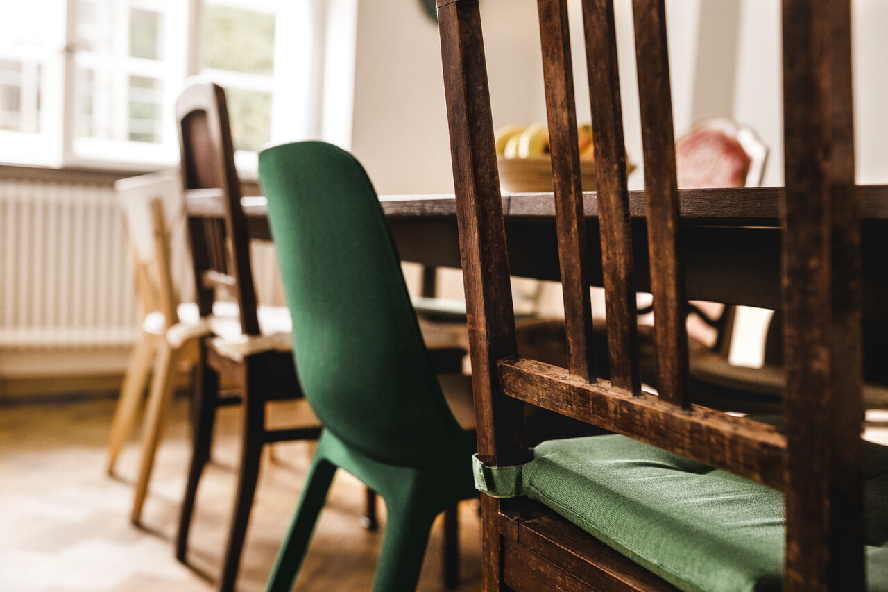 Blick durch die Lehne eines alten Holzstuhls auf den langen Esstisch im POSTYOU-Büro. Um den Tisch stehen unterschiedliche Stühle, darunter ein grüner Schalenstuhl; im Hintergrund helle Fenster.
