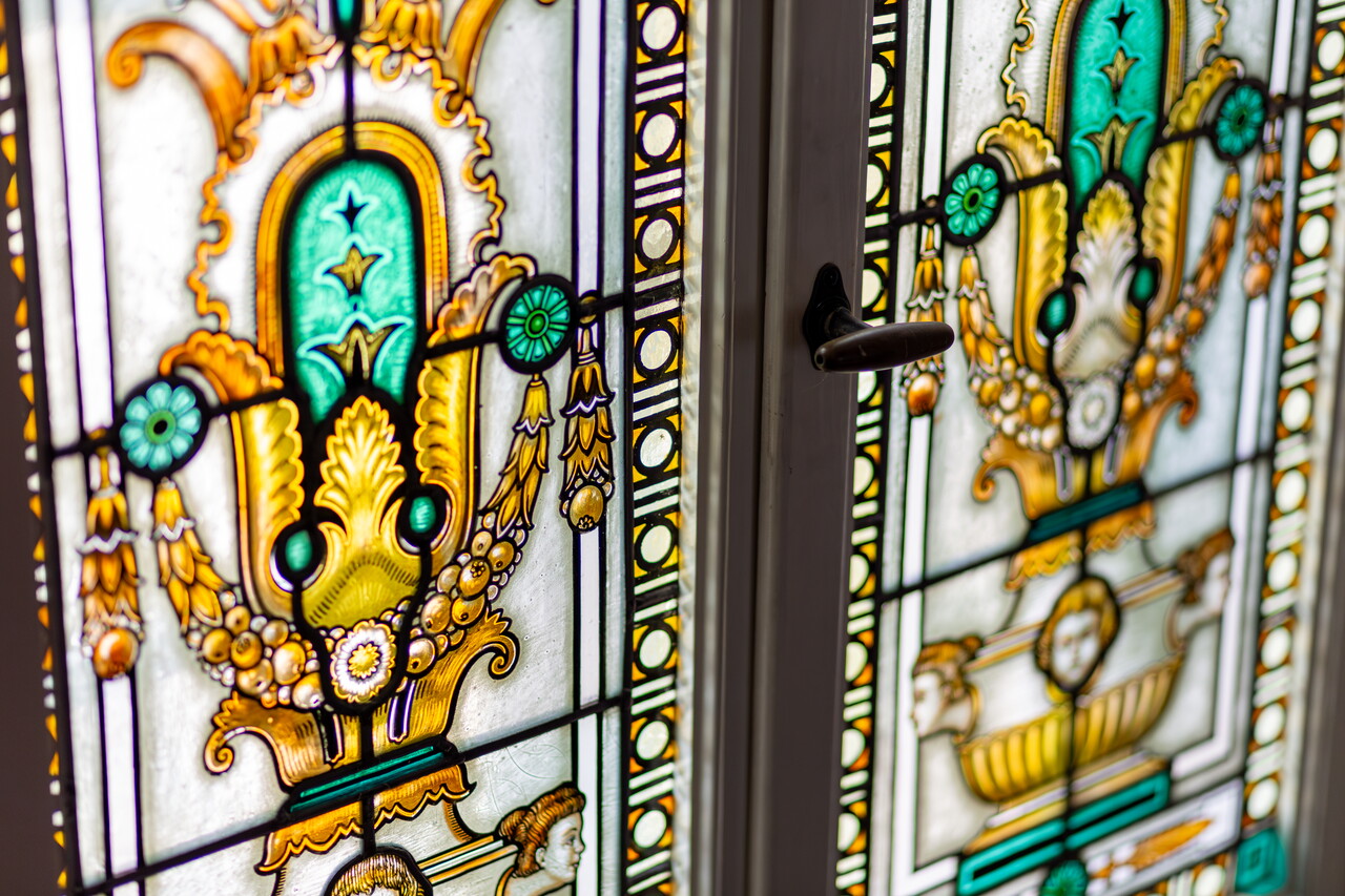 Nahaufnahme eines farbigen Bleiglasfensters in der Villa der Agentur. Die Ornamente zeigen gelbe, grüne und türkise Muster mit Ranken und Vasenformen.