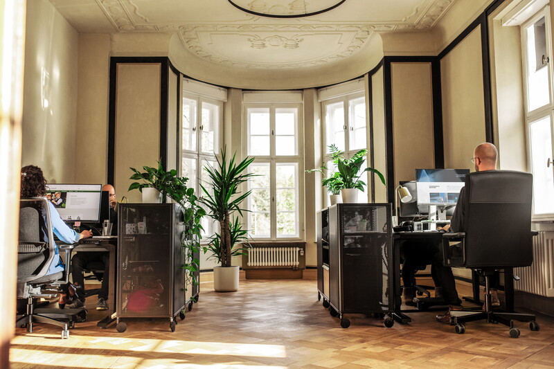Ein heller Altbauraum im POSTYOU-Büro mit Stuckdecke, hohen Fenstern und Parkett. Im Erker stehen schwarze Schreibtische mit Monitoren und Pflanzen, an einem davon arbeitet ein Mitarbeiter des POSTYOU-Teams.