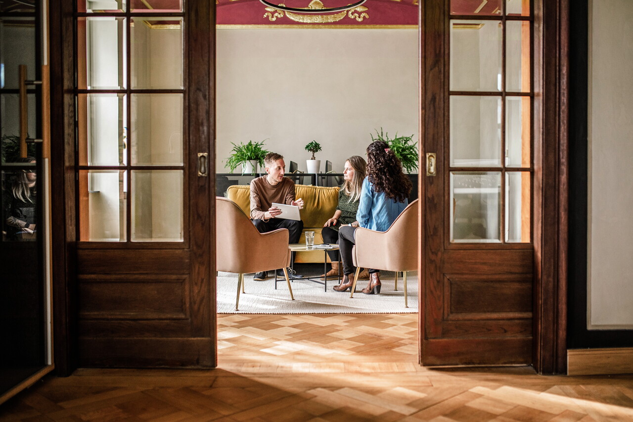 Blick durch eine geöffnete, dunkle Holzflügeltür in den Besprechungsraum von POSTYOU mit roter Stuckdecke. Dort sitzen mehrere Mitglieder des POSTYOU-Teams auf einem gelben Sofa und in Sesseln.
