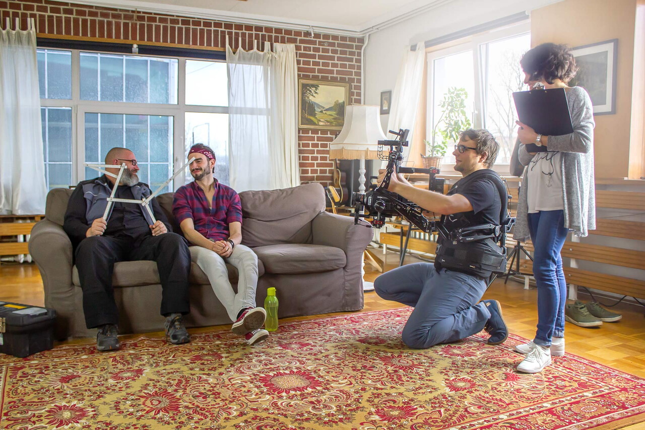 Videodreh in einem Wohnzimmer mit Backsteinwand und Perserteppich: Zwei Männer sitzen auf einem Sofa, ein Kameramann kniet mit Gimbal davor. Eine Kollegin schirmt mit einem Reflektor das Licht ab.