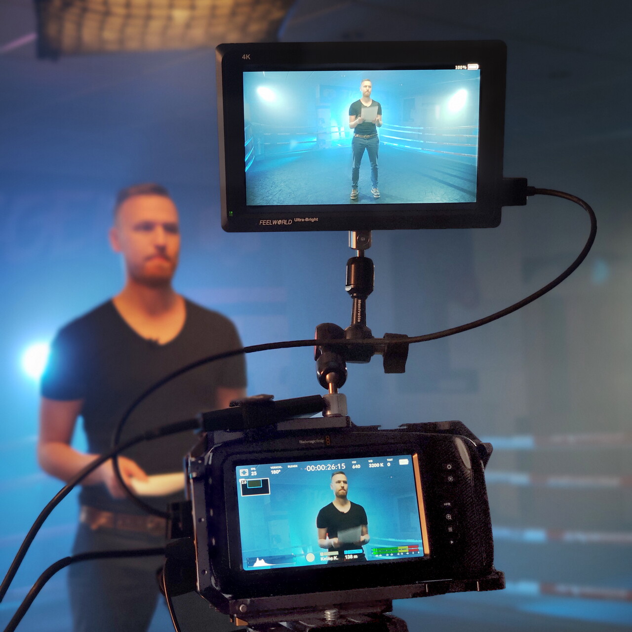 Interviewdreh in einem blau beleuchteten Studio: Im Vordergrund stehen eine Kamera und ein Kontrollmonitor, die beide den Sprecher zeigen. Dahinter steht der Mann unscharf vor einer Leuchte.
