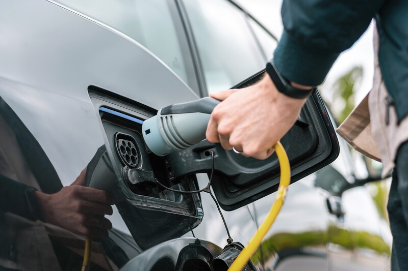 Eine Hand steckt einen Ladestecker mit gelbem Kabel in die Ladebuchse eines silbernen Elektroautos.