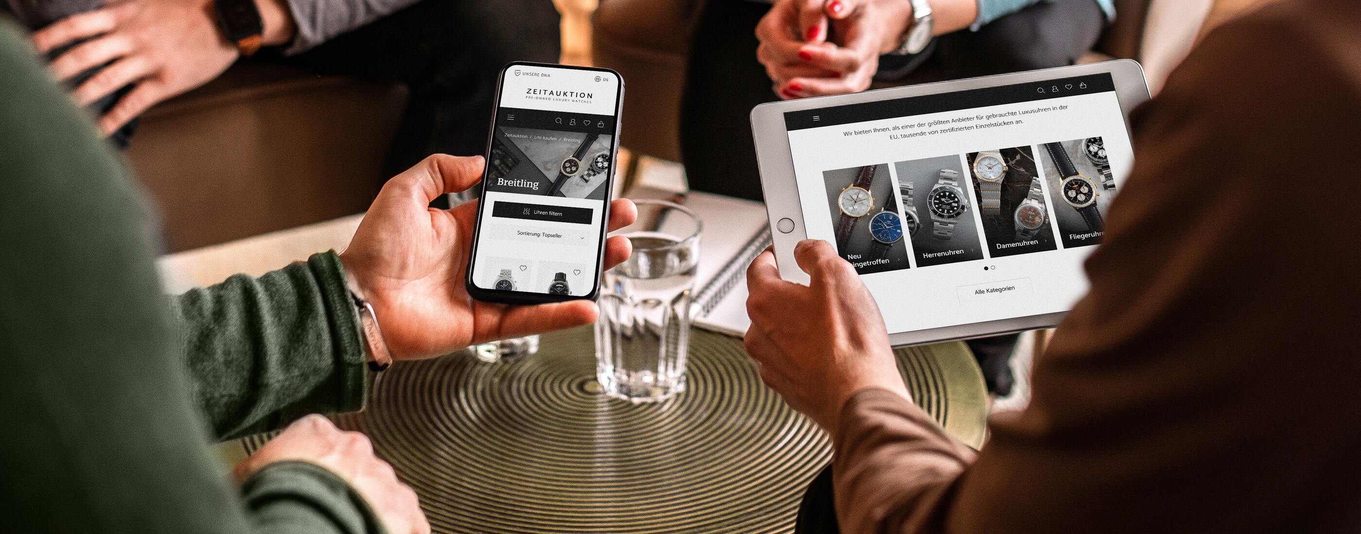 Nahaufnahme einer Gesprächsrunde im POSTYOU-Team: Eine Hand hält ein Smartphone, eine andere ein Tablet, beide zeigen einen Onlineshop für Uhren. Dazwischen steht ein Wasserglas auf einem goldenen Tisch.