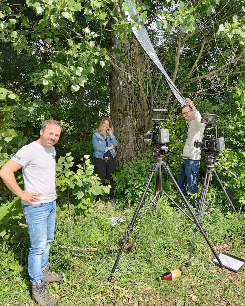Außendreh im Grünen: Zwei Kameras auf Stativen sind auf eine Frau vor einem großen Baum gerichtet. Ein Mann hält eine Tonangel, ein anderer steht lächelnd im Vordergrund.